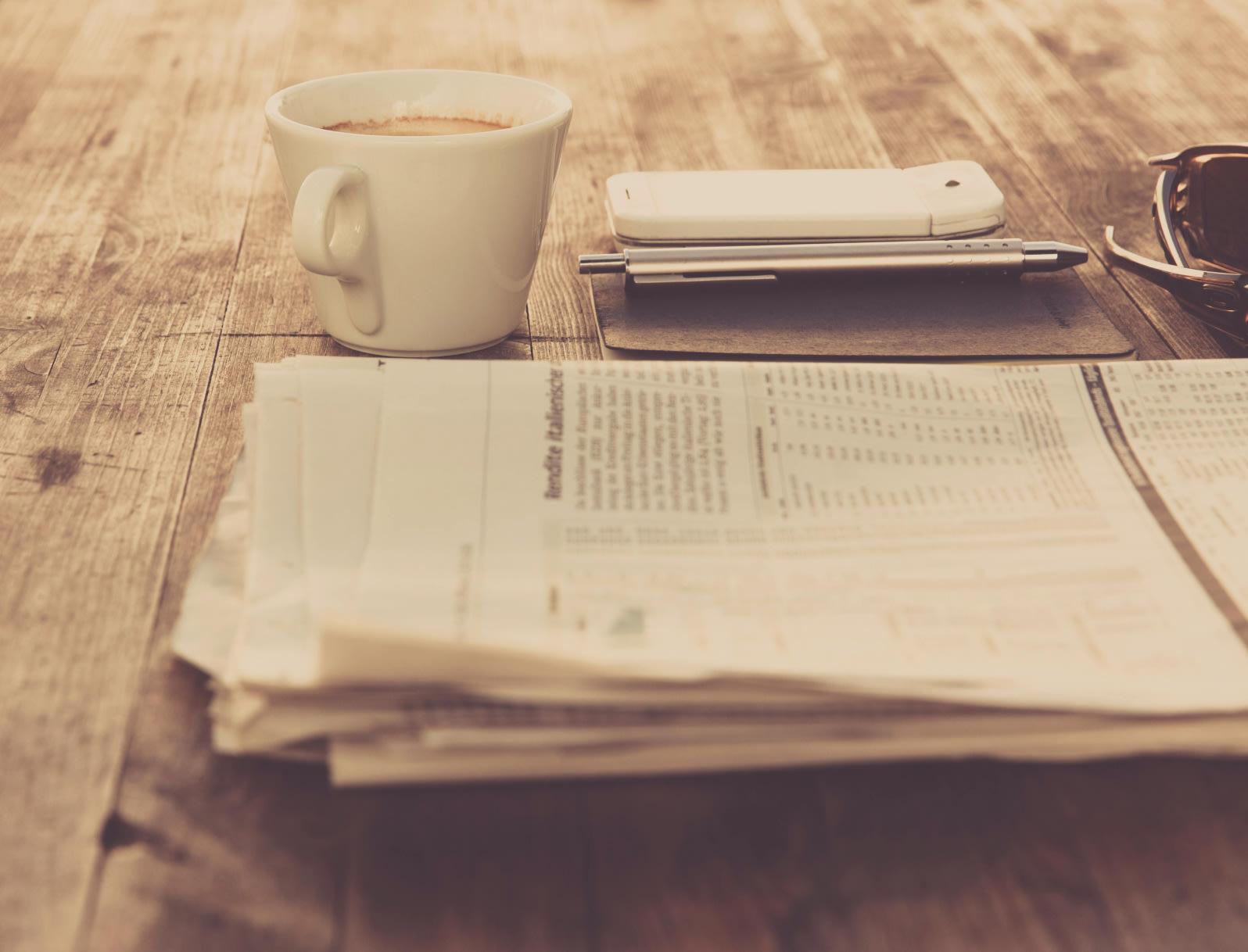 Eine Zeitung liegt auf einem Tisch. Daneben eine Tasse Kaffee und weitere Schreibutensilien